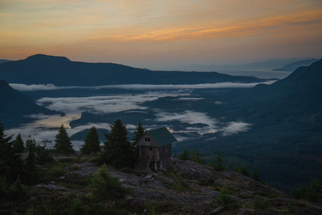 A sunset scene of a cabin overlooking a low cloud hanging valley, with mountians in the background.