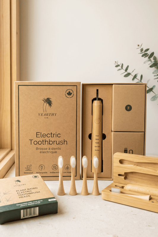A bamboo electric toothbrush starter kit from Vearthy sits on a brightly lit white counter by a window. Included are the 4 replacement heads, handle with 2 stock replacement heads and the travel case made from bamboo. 