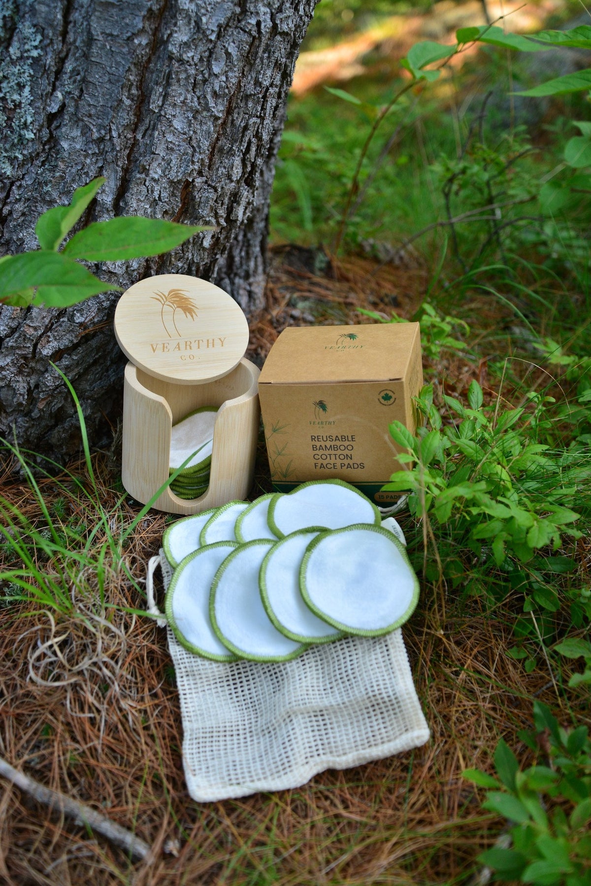 A set of reusable bamboo face pads with and matching cloth bags arranged outdoors on a tree trunk with pine needles and green plants in the background. The set includes a small bamboo holder,  a kraft paper box, and 15 round reusable cloth face pads with white interiors and green edges. 